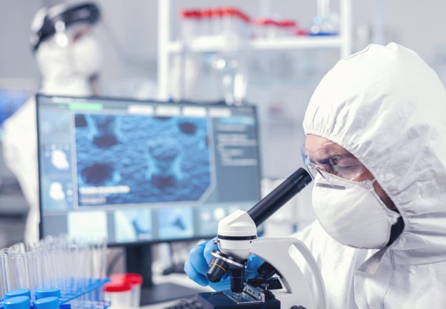Medical team in laboratory conducting coronavirus analysis using microscope. Scientist in protective suit sitting at workplace using modern medical technology during global epidemic.